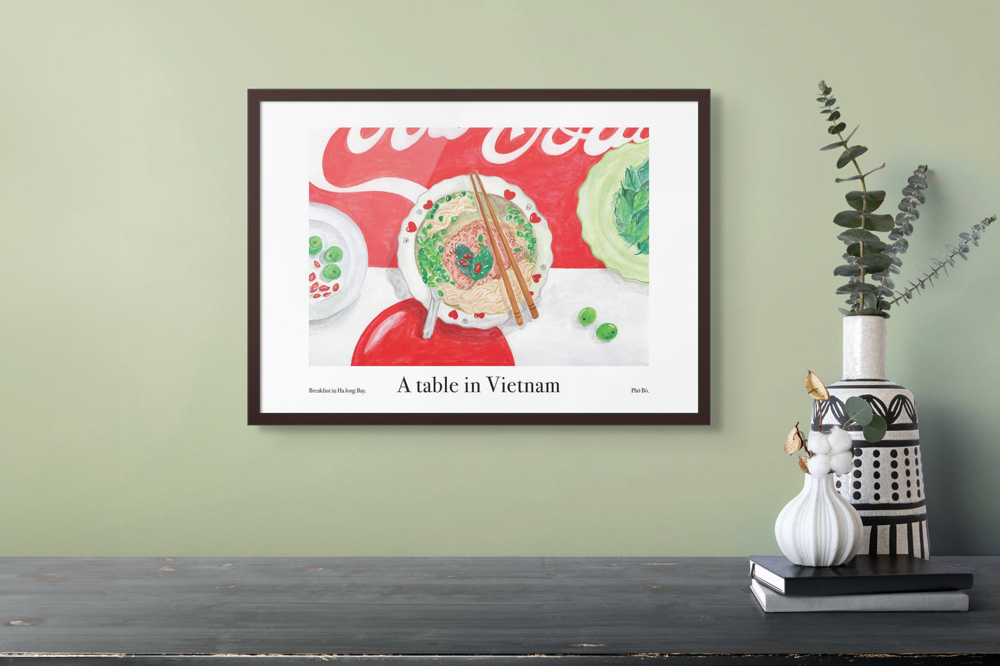A room with an art poster on the wall of a table in Vietnam with a bowl of Pho.