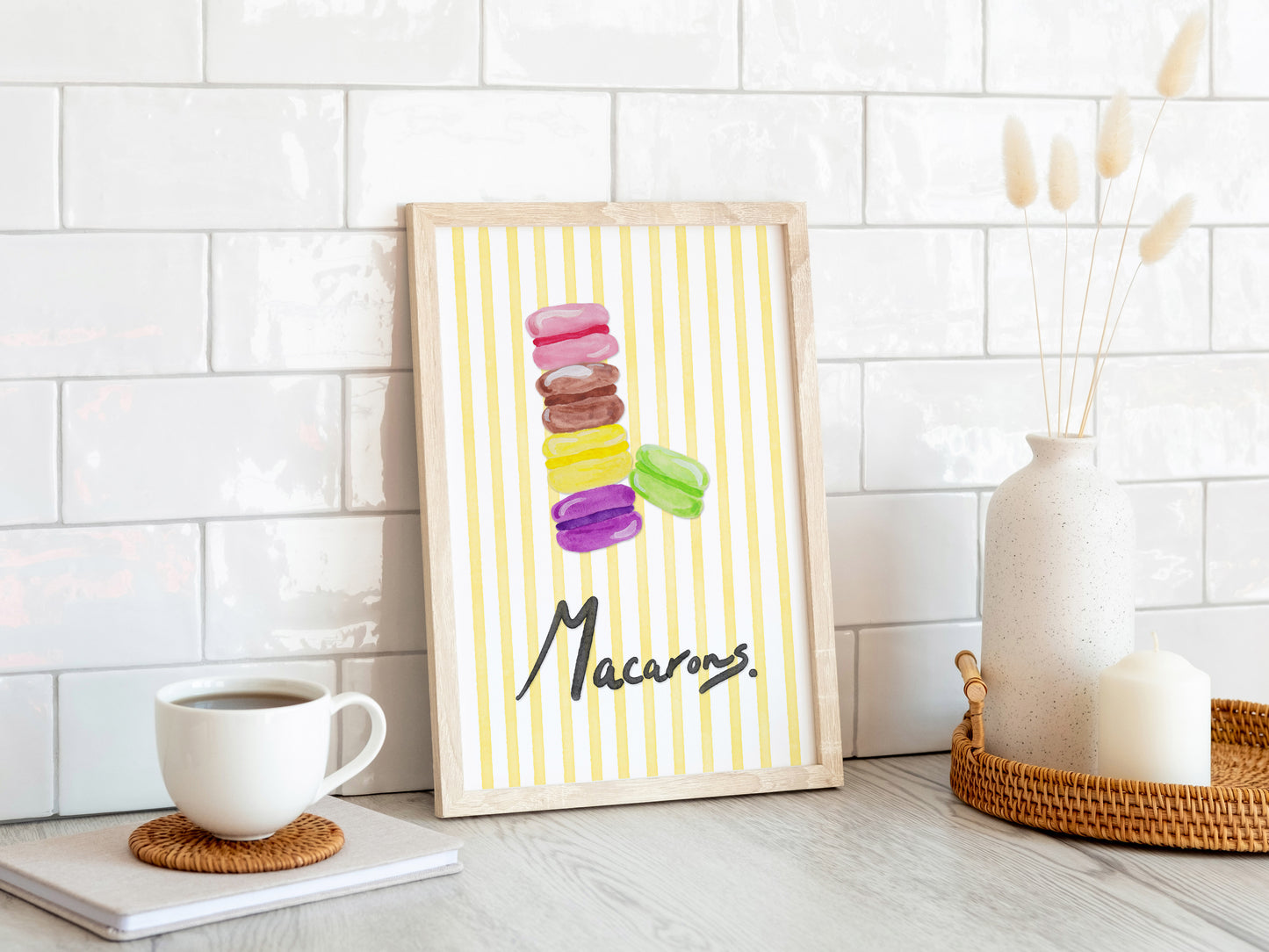 Framed artwork of colorful macarons on a white wall with a coffee cup and decor items.