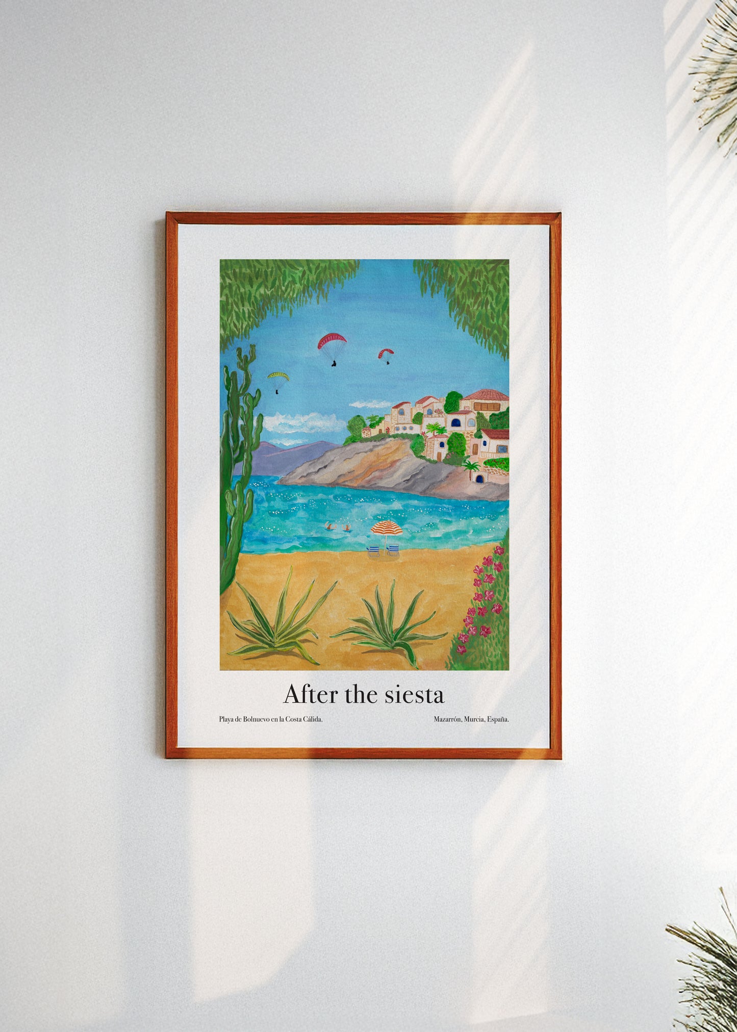 Framed artwork of a coastal scene in Spain with houses and paragliders, titled 'After the siesta', on a white wall.