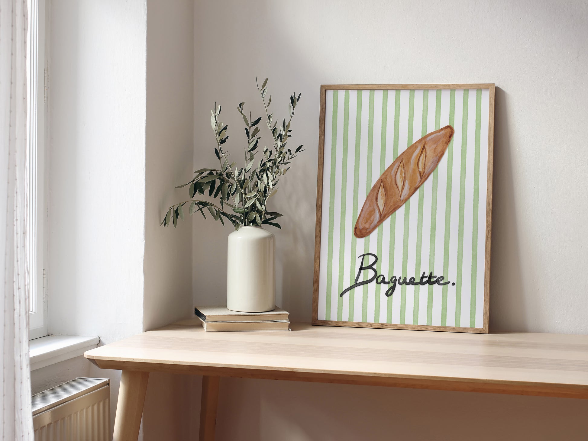 Framed artwork of a baguette on a striped background, placed on a wooden surface with a vase and books.