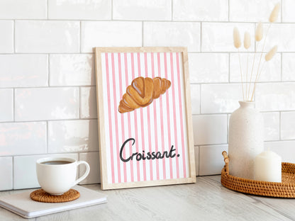 Framed artwork with a croissant illustration on a pink striped background, placed on a white surface with a coffee cup and decor items.
