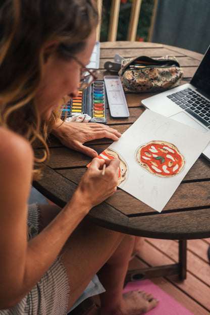 Woman drawing a pizza on paper with a laptop and art supplies on a wooden table.