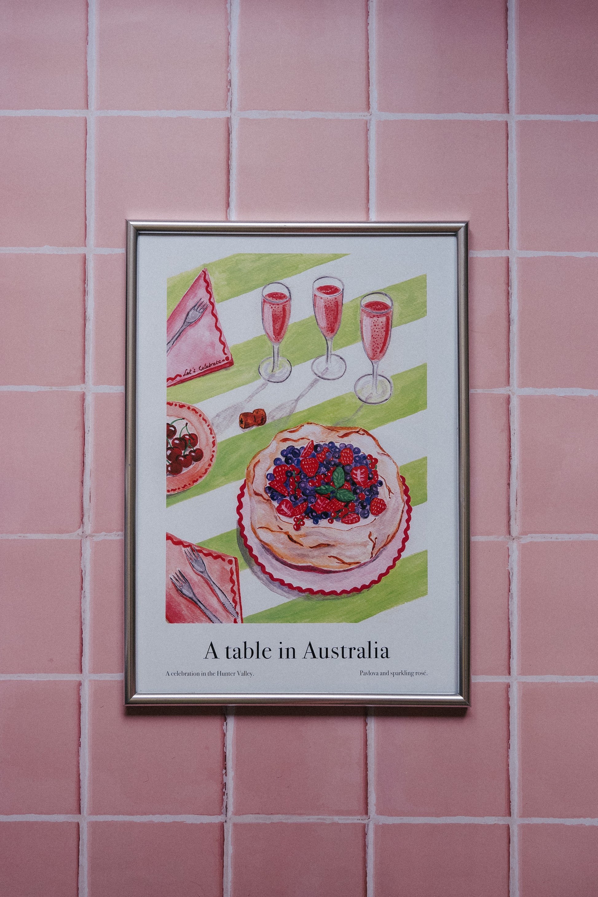 Framed artwork of a table setting with food and drinks on a pink tiled wall.