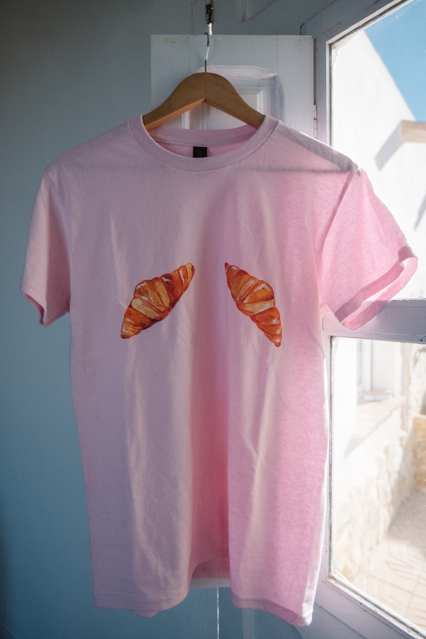 A pink t-shirt with hand-painted croissants hanging on a coat hanger