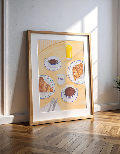 Framed artwork of a breakfast scene with croissants and coffee on a yellow background, leaning against a white wall.